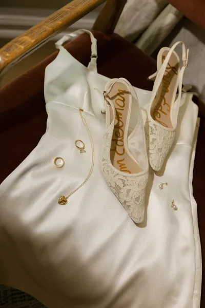 Wedding shoes with lace wedding heels arranged on a satin slip dress with gold heart pendant, earrings, and ring on a wooden chair