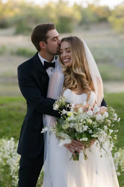 Couple portrait of groom kissing bride’s forehead as she holds a white and green bouquet, long veil flowing in a wildflower field