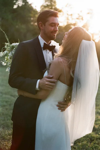 Couple portrait of bride and groom embrace at sunset, her long veil and bouquet visible as golden light filters through trees on a lawn