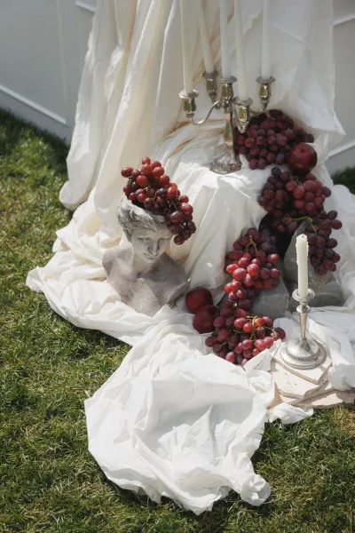 Wedding decor vignette with styled wedding details, white draped fabric, silver candelabra, taper candles, and grapes on a lawn backdrop