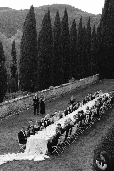 Outdoor wedding reception with a long wedding banquet table set in white linens, candles and place settings as black-tie guests dine on a lawn by a stone wall and cypress trees