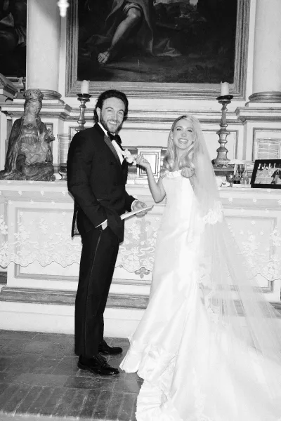 Wedding couple portrait in black and white, bride in veil and strapless dress holds groom’s lapel by a candlelit fireplace mantel indoors