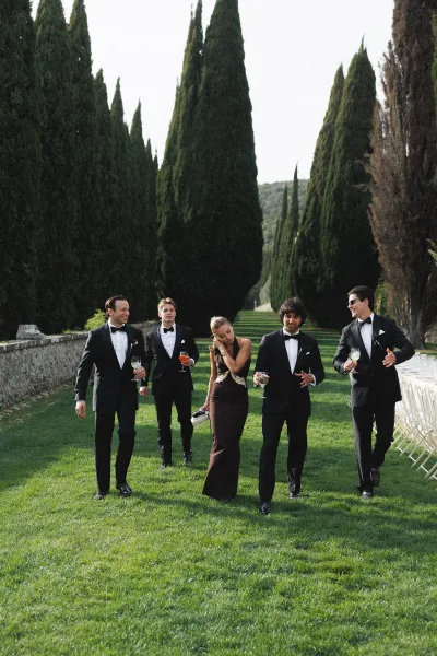Wedding party photo of groomsmen walking with a bridesmaid, holding cocktails in black tuxedos and sunglasses on a lawn by cypress trees
