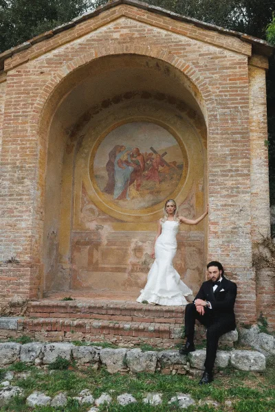 Couple portrait of bride in a strapless satin mermaid gown and groom in a black tuxedo under a brick archway with fresco mural backdrop