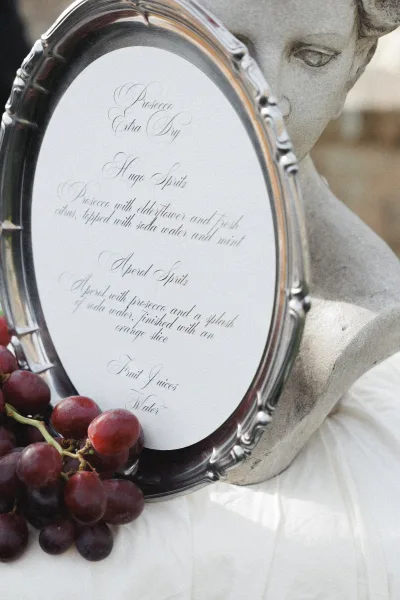 Wedding bar menu in an ornate silver frame with calligraphy and grape accents on a white tablecloth in a neutral outdoor setting