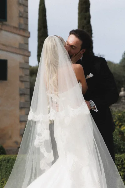 Wedding kiss as bride and groom embrace under a lace-trim veil, tuxedo pocket square and watch visible by stone building and cypress trees
