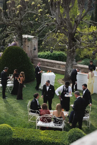 Cocktail hour with wedding guests mingling around a garden cocktail hour bar, bartender pouring drinks beside white linen high tops and iron chairs