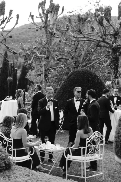 Wedding cocktail hour with guests in tuxedos and sunglasses mingling around white linen tables on a garden lawn with distant hills