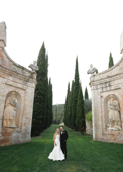 Couple portrait of bride and groom portrait in a garden, she wears a veil by brick stone walls with statues and cypress trees behind