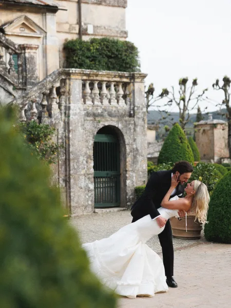 Couple portrait of groom dipping bride in a playful wedding dip pose, her strapless dress flowing in a stone courtyard garden by stairs