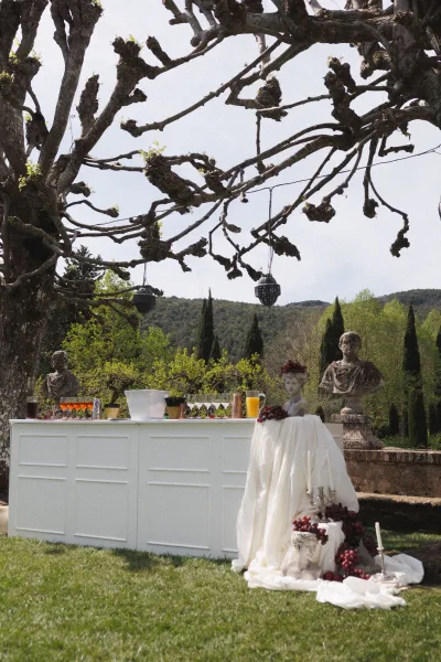 Outdoor wedding bar with a white counter, cocktail glasses and pitchers, taper candle candelabra, and lanterns hanging from tree branches on a stone terrace