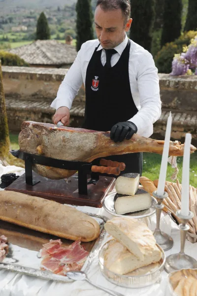 Wedding catering station with a prosciutto leg on a carving stand, bread and cheese on silver trays, set on an outdoor stone terrace