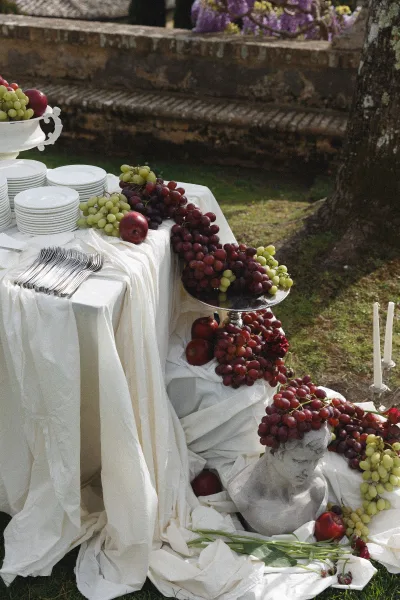 Wedding buffet table with wedding fruit display of grapes and apples, white draping, plates and silver cutlery, taper candles by stone steps outdoors