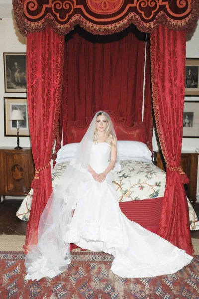 Bridal portrait of a bride sitting on bed, veil draped over a strapless tulle gown on a canopy bed with red velvet curtains behind her