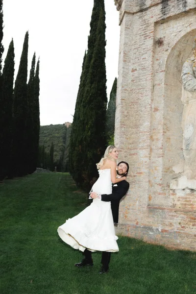 Couple portrait of groom lifting bride as they laugh, her strapless dress and his bow tie by a stone wall and brick arch outdoors