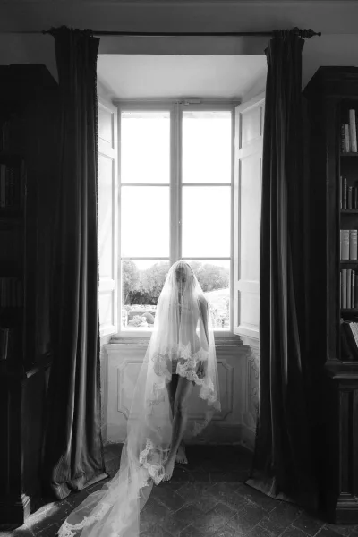 Bridal portrait of a bride in veil with a lace veil draped over her face, standing by a tall window in a book-lined room