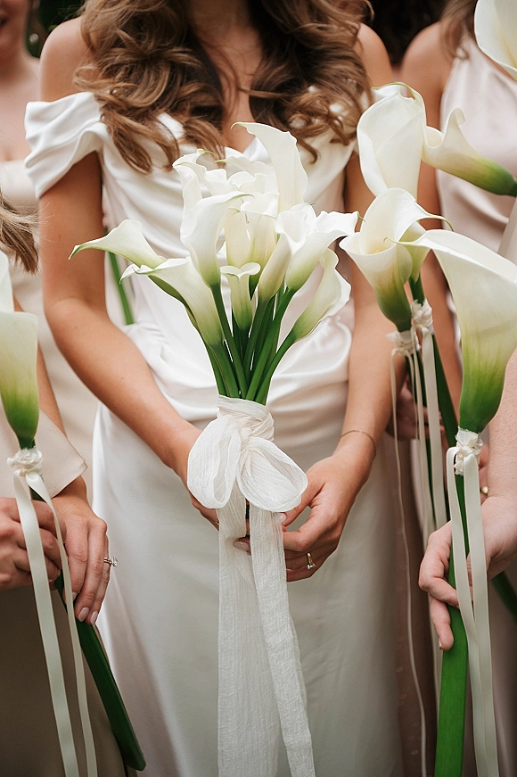 Bridesmaid bouquet of white calla lilies with trailing ribbon, held by bridesmaids in satin off-shoulder dresses with rings in hand