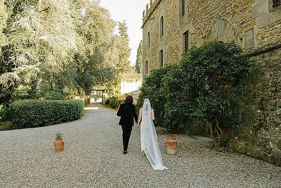 Couple portrait of newlyweds walking away holding hands, bride’s long lace veil and dress train beside terracotta pots in a stone courtyard