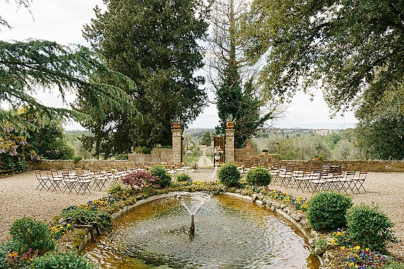 Outdoor ceremony setup with wood folding chairs lining a gravel aisle, floral arrangements by stone pillars, and a garden fountain in back