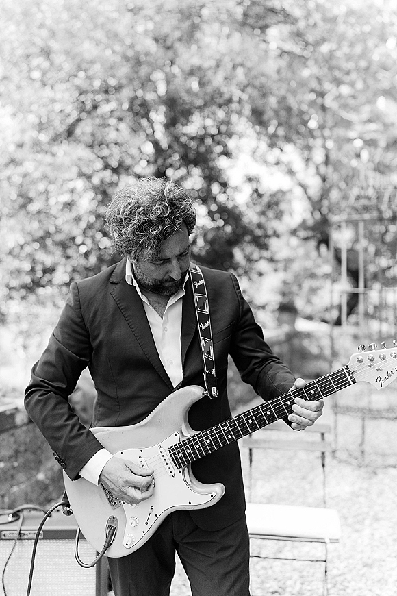 Wedding guitarist playing electric guitar in a suit jacket beside an amplifier, framed by soft garden foliage bokeh in the background