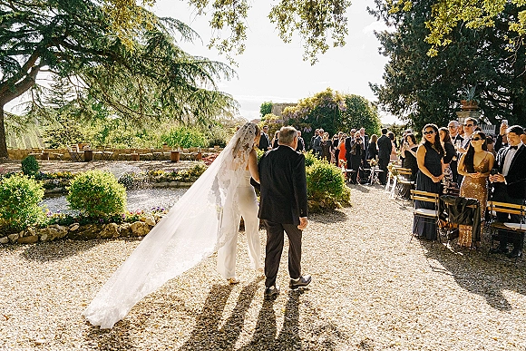 Wedding processional as bride walking down aisle with her father, lace veil train trailing on a gravel path past standing guests in a garden near a pond