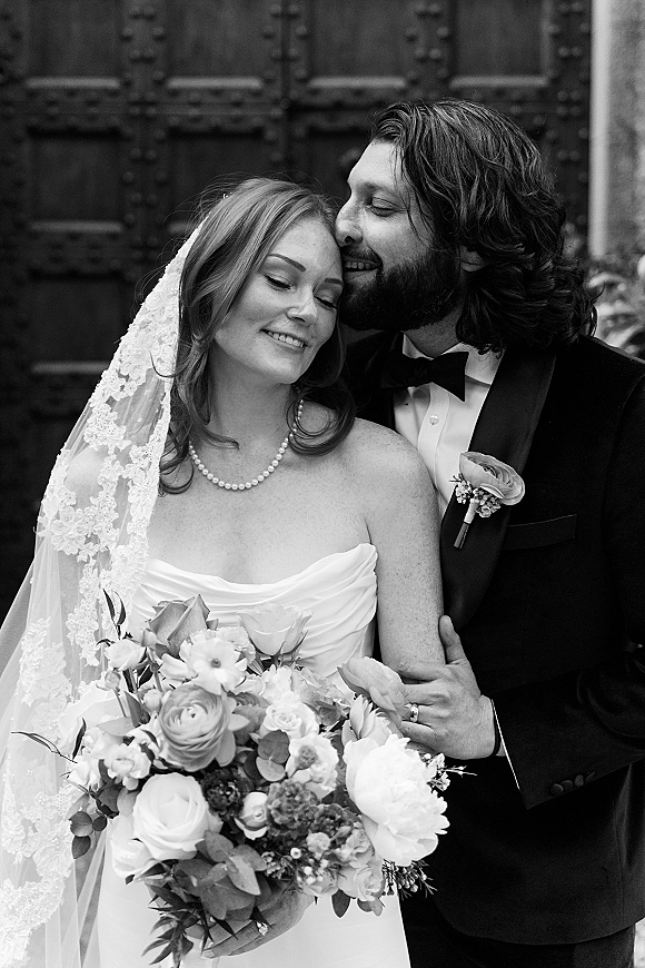 Couple portrait, black and white wedding portrait of groom kissing bride’s forehead as she holds a rose bouquet before large wooden doors