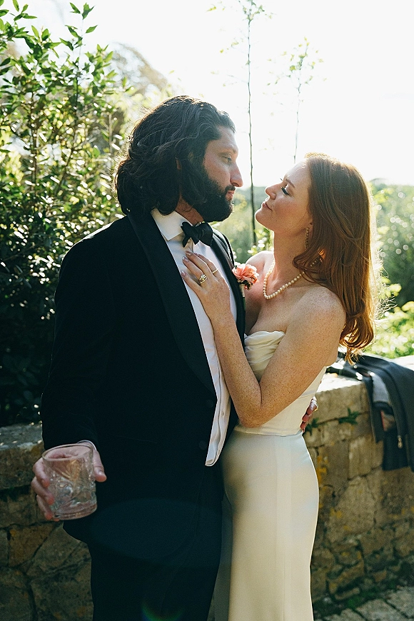Couple portrait of bride and groom embrace, her hand on his chest as he holds a whiskey glass by a sunlit stone wall in lush garden greenery