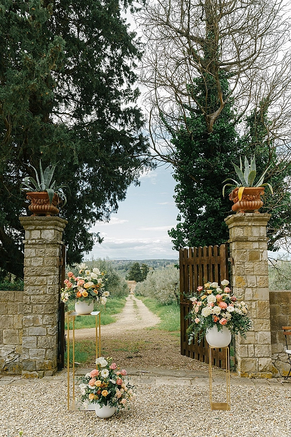 Ceremony entrance decor with wedding entrance flowers in white ceramic urns on gold stands framing a stone gate and gravel courtyard