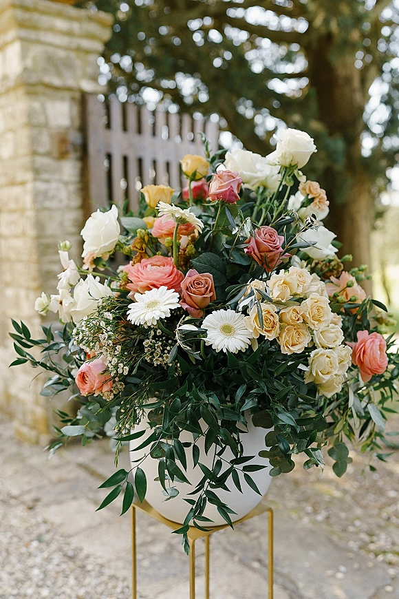 Wedding floral arrangement with roses and pastel blooms in a white urn on a gold pedestal beside a stone pillar and picket fence outdoors