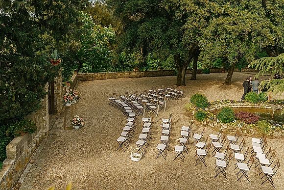 Outdoor ceremony setup with folding chairs and floral arrangements lining a gravel aisle in a stone-walled garden courtyard with trees