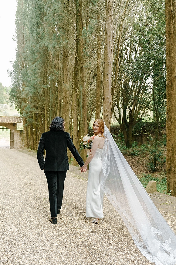 Couple portrait of bride and groom walking away hand in hand, bride looking back with lace veil and bouquet on a tree-lined path