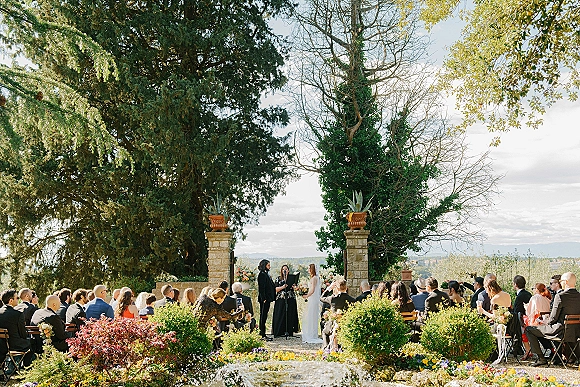 Outdoor wedding ceremony with floral arch and guests seated in wooden chairs along a gravel aisle between stone pillars and trees