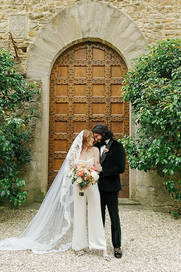 Couple portrait of bride and groom embracing with bouquet and lace veil by a stone wall and arched wooden door in courtyard