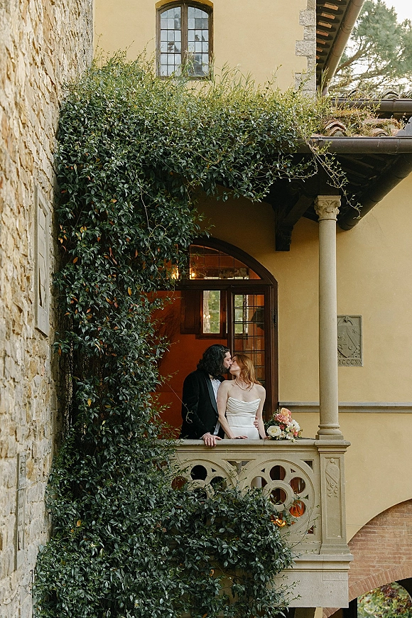 Wedding couple portrait of bride in a strapless dress and groom in a suit kissing on a balcony, holding bouquet against an ivy-covered stone villa