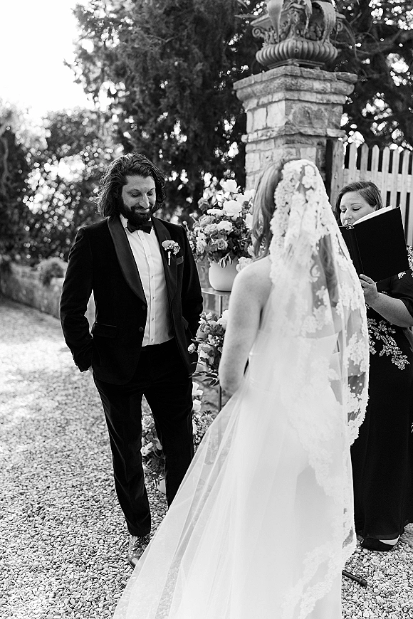 Wedding vows during an outdoor wedding ceremony as bride in lace veil faces groom in tux beside stone pillar and floral pedestal