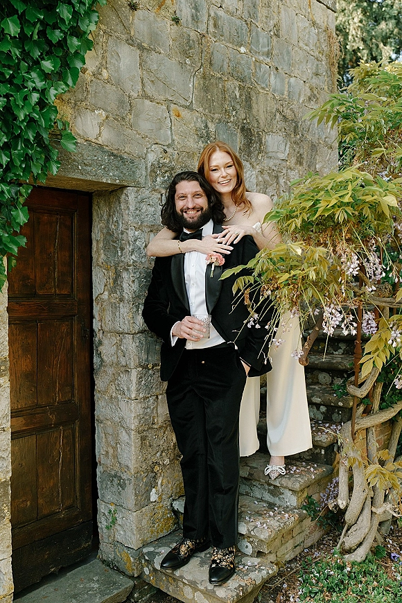 Couple portrait of bride hugging groom in tuxedo, her pearl necklace and strapless dress framed by ivy on stone steps by a wooden door