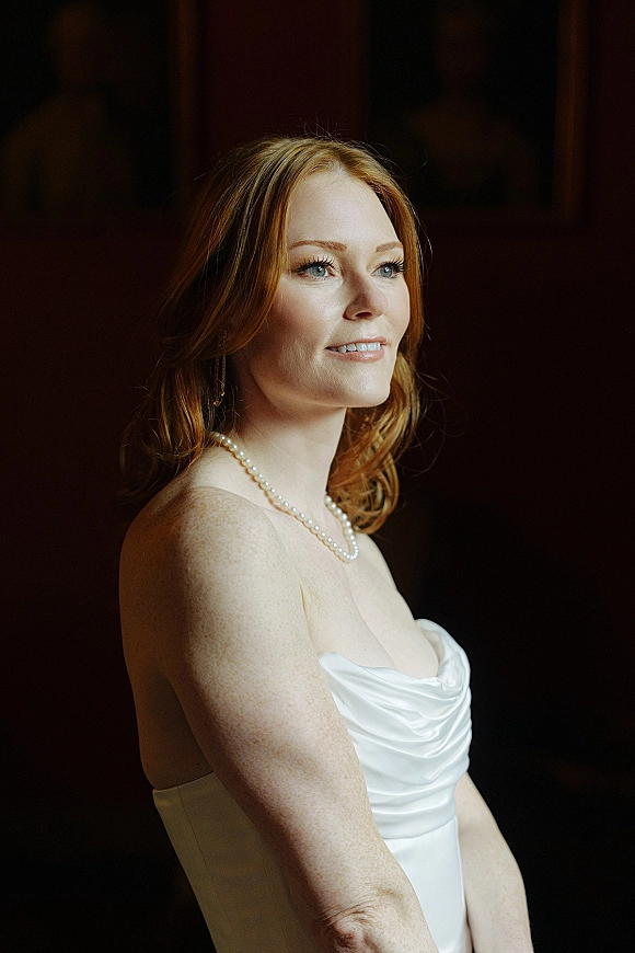 Bridal portrait of a bride in side profile wearing a strapless satin wedding dress and pearl necklace against a dark indoor backdrop