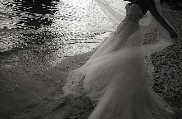 Bridal portrait of a bride on beach in a strapless tulle wedding dress, long veil trailing behind her by the shoreline and ocean water