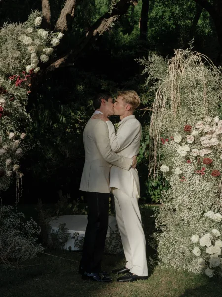 Wedding kiss between two grooms in light-colored suits under a white rose floral arch with hanging strands on a garden lawn