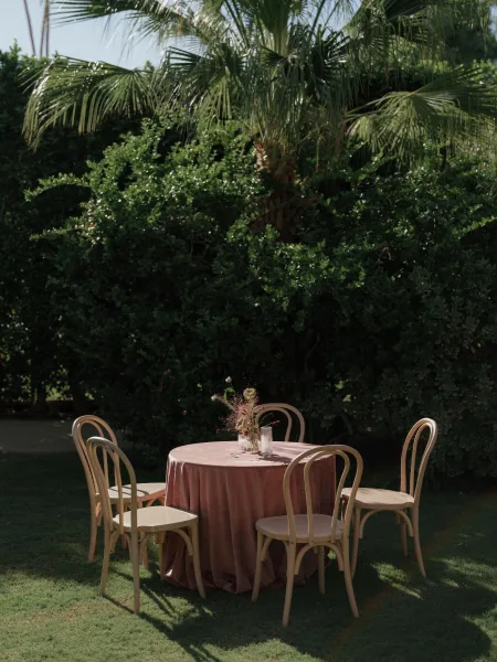 Reception tablescape with mauve tablecloth on a round outdoor reception table, bud vase dried florals and taper candles, palm garden backdrop
