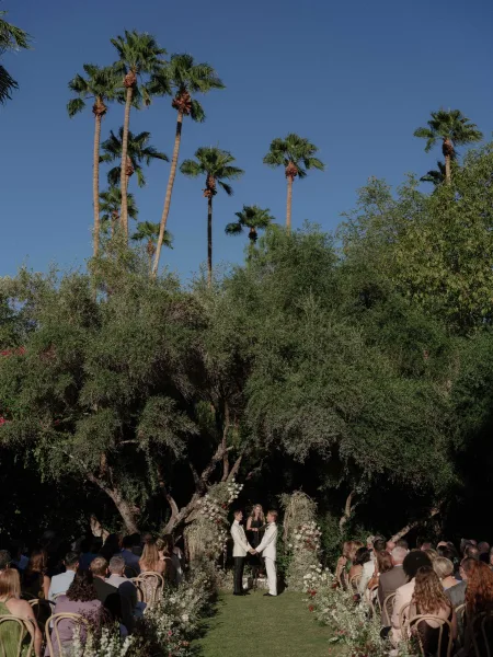 Ceremony moment at an outdoor wedding ceremony with two grooms holding hands beneath a floral arch, guests seated on a lawn under palm trees