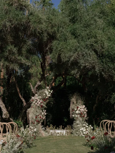 Ceremony setup with an outdoor ceremony setup of wooden chairs facing hanging roses and greenery, with a white aisle runner on lawn under trees
