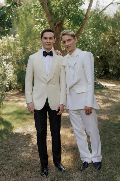 Groom portrait in a white tuxedo jacket with black bow tie and wedding ring, standing in dappled sunlight among garden trees