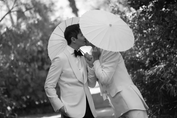 Wedding kiss portrait of one groom kissing the other beneath a paper parasol, both in white tuxedos on a tree-lined garden path