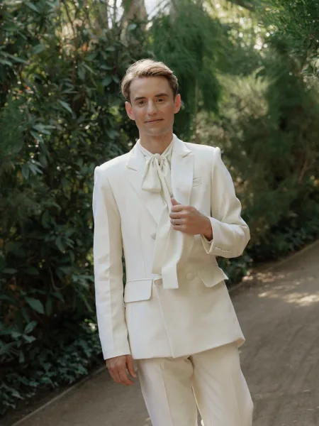 Groom portrait in a white tuxedo with bow tie and lapel buttons, wedding band visible, standing on an outdoor garden path among trees