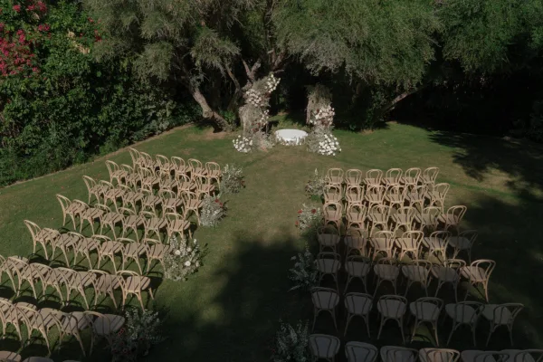 Outdoor ceremony setup with curved chairs facing a floral arch on a white round platform, aisle flowers on a sunny garden lawn