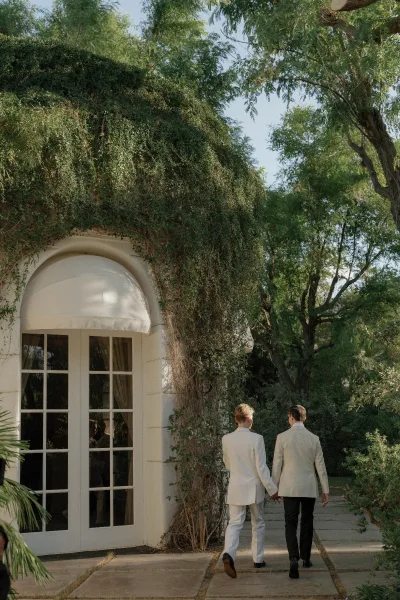 Couple portrait of grooms holding hands in white suits with boutonnieres, walking past an ivy-covered building with arched window and French doors