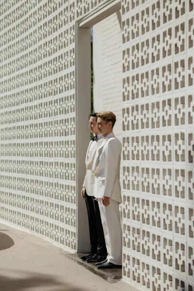 Groom portrait of two grooms in tuxedos, one in white and one in black with bow ties, standing by a patterned concrete wall in sun shadows