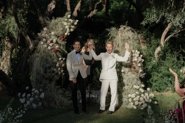 Recessional moment as two grooms walk the aisle, raising hands in celebration beneath a floral arch of white roses and baby's breath outdoors