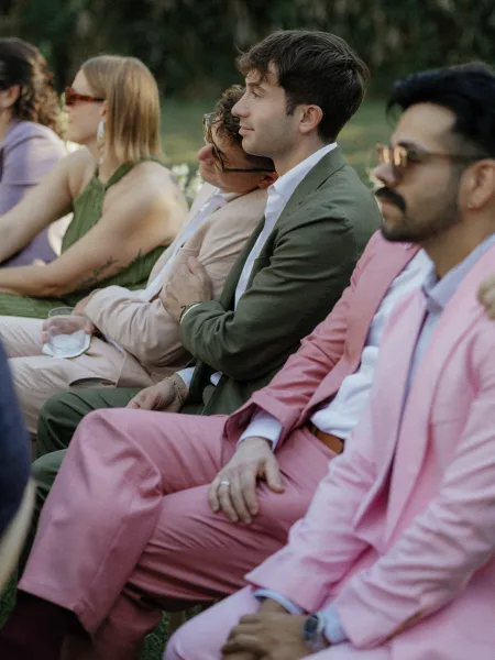 Wedding guests in pink suits and sunglasses seated on ceremony chairs, one holding a drink, on an outdoor lawn with trees